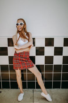 Fashionable woman in checkered skirt poses playfully against a retro wall with sunglasses.