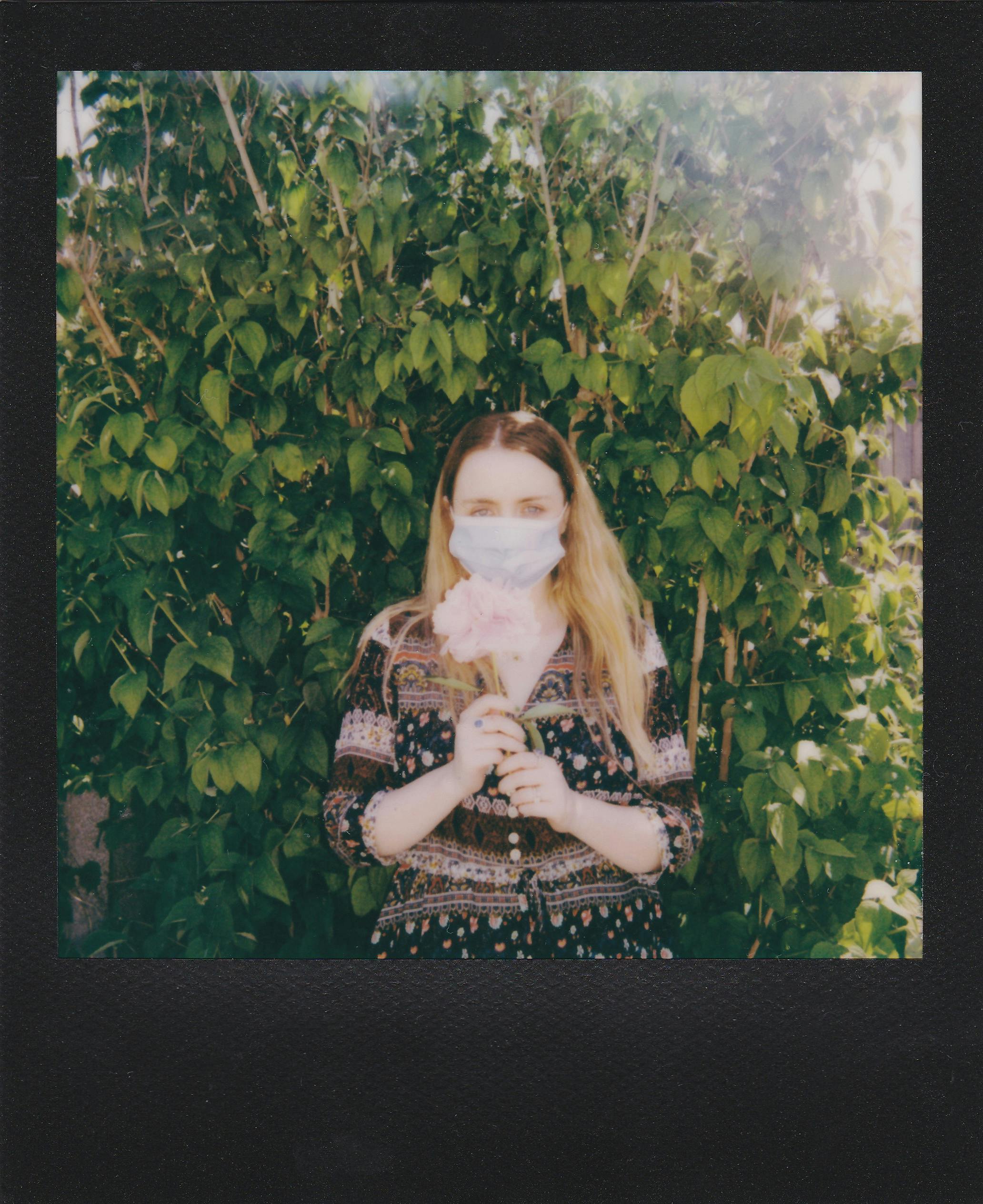 A woman wearing a face mask holds a pink flower against a backdrop of lush green plants.