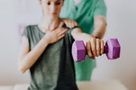 在医院的医生办公室里，一名面目全非的骨科医生穿着制服，帮助一位穿着休闲装的苗条女性用哑铃伸展手臂