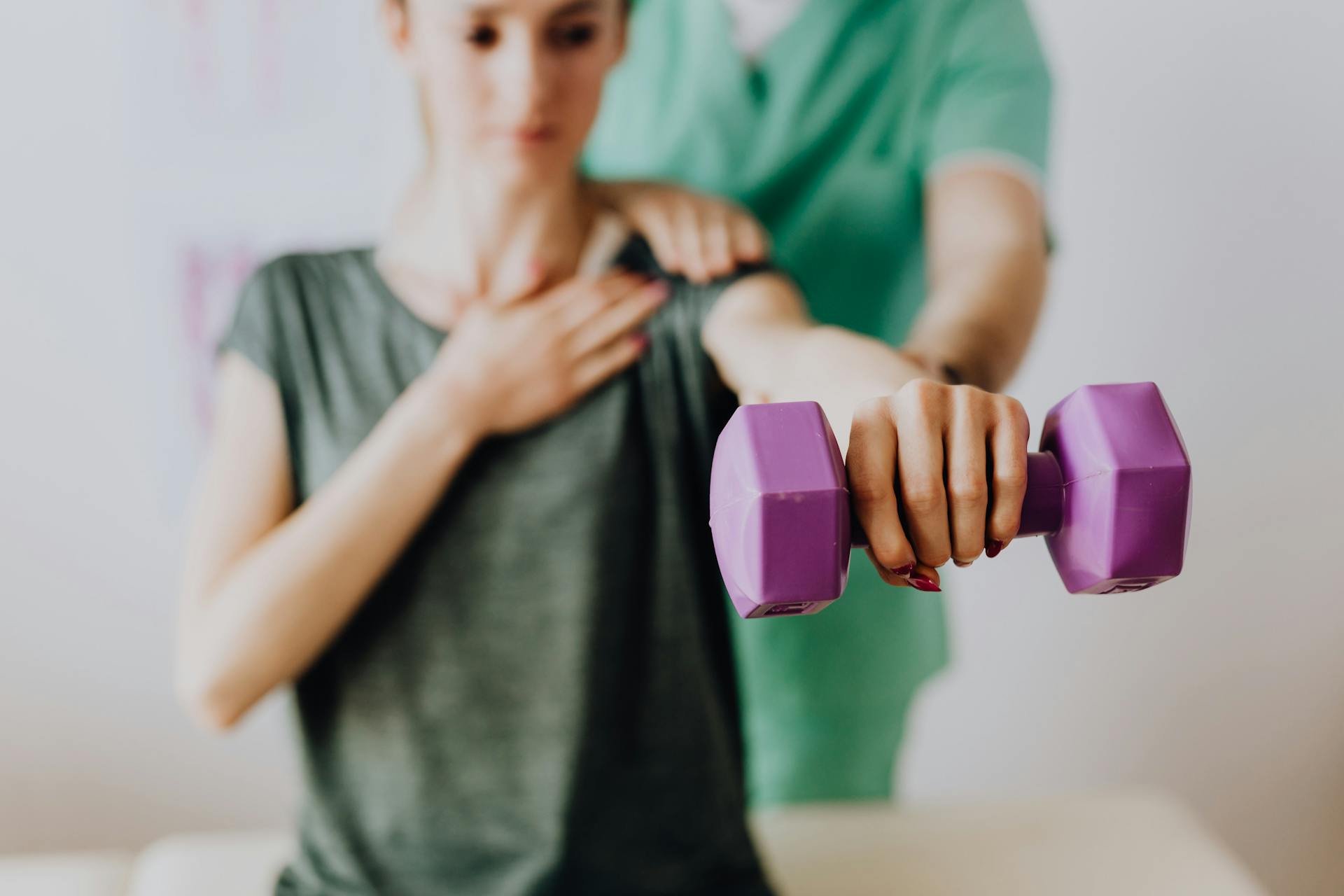 Crop unrecognizable orthopedist in uniform helping slender female in casual wear reaching arm with dumbbell in doctor office in hospital