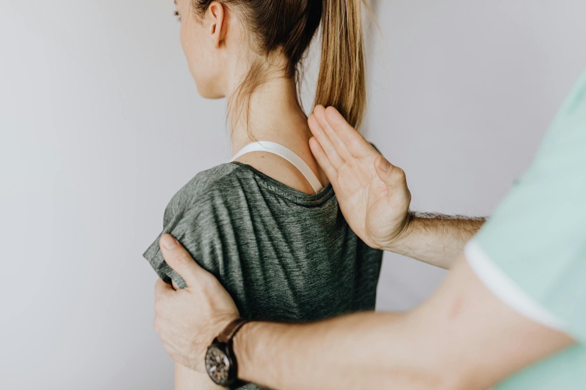 Back view of crop unrecognizable osteopath in uniform and wristwatch checking up back of slim female patient in casual wear on white background