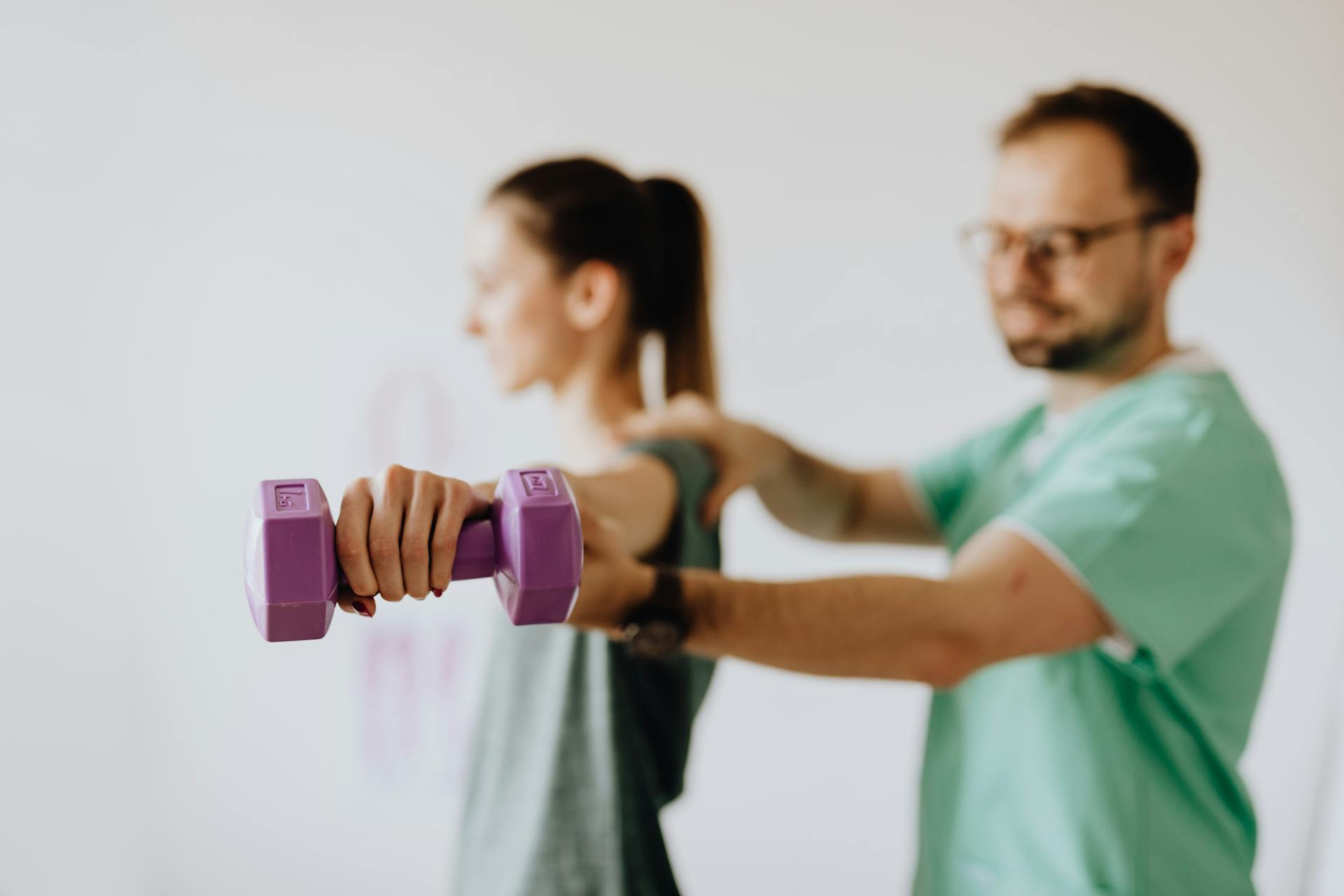 Side view of professional orthopedist in uniform and eyewear helping fit woman reaching arm with dumbbell in doctor office on blurred background