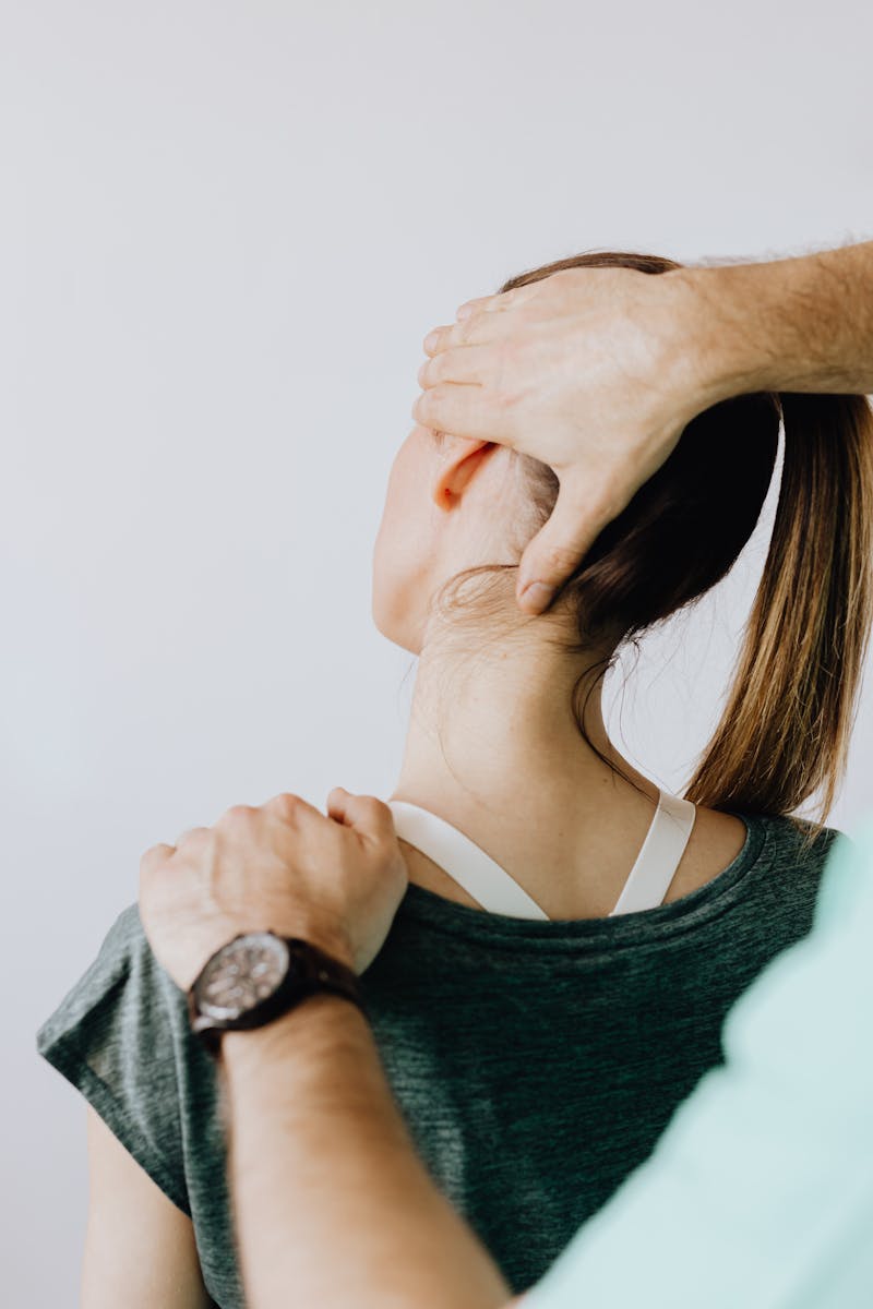 Back view of crop faceless chiropractor in wristwatch stretching neck of unrecognizable female patient during medical examination in doctor office in clinic