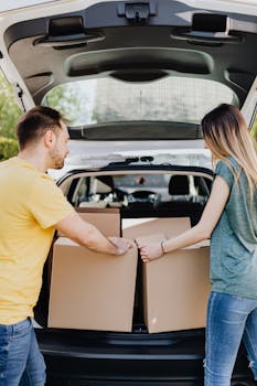 A young couple loads cardboard boxes into their car trunk, preparing for a move on a sunny day.