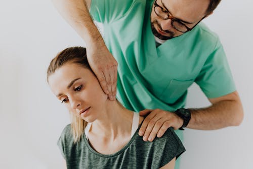 Confident doctor wearing uniform and eyeglasses gently doing therapeutic massage on calm female patients neck and stretching stiff neck muscles