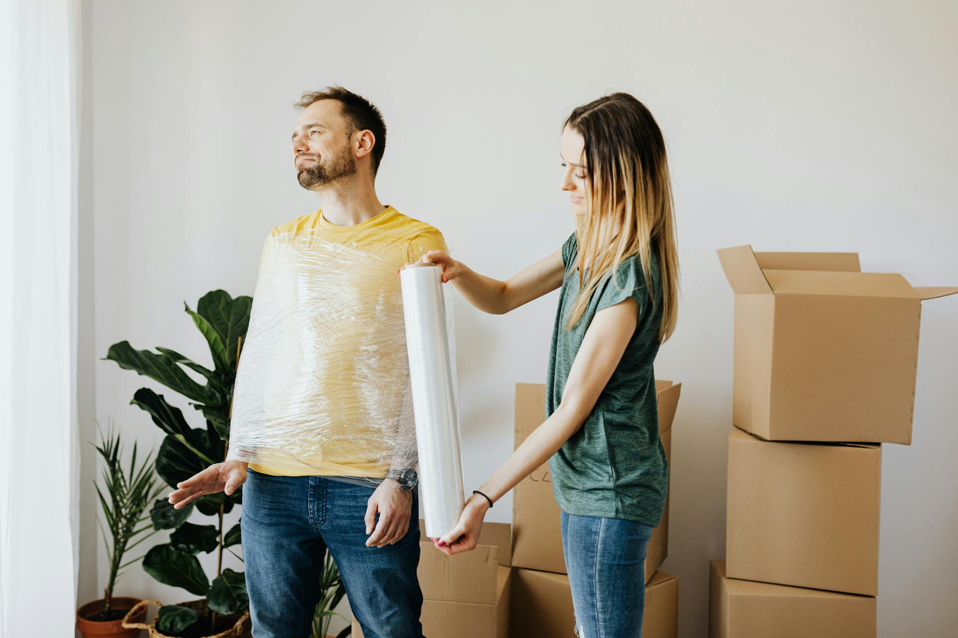 Content couple having fun while girlfriend in casual wear wrapping boyfriend with clingy plastic roll during packing belongings into carton boxes before moving in together