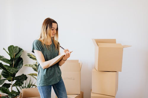 Focused young lady in casual wear taking notes in clipboard while standing near packed carton boxes before moving into new house