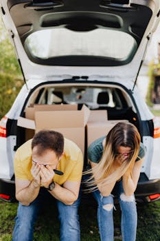 A distressed couple sits by a car full of boxes, symbolizing moving stress.