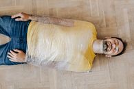 Top view young bearded male in casual outfit lying with eyes closed on floor with upper body wrapped by using clingy plastic