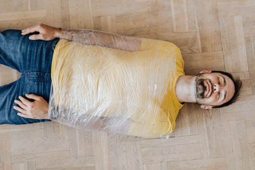 Top view young bearded male in casual outfit lying with eyes closed on floor with upper body wrapped by using clingy plastic