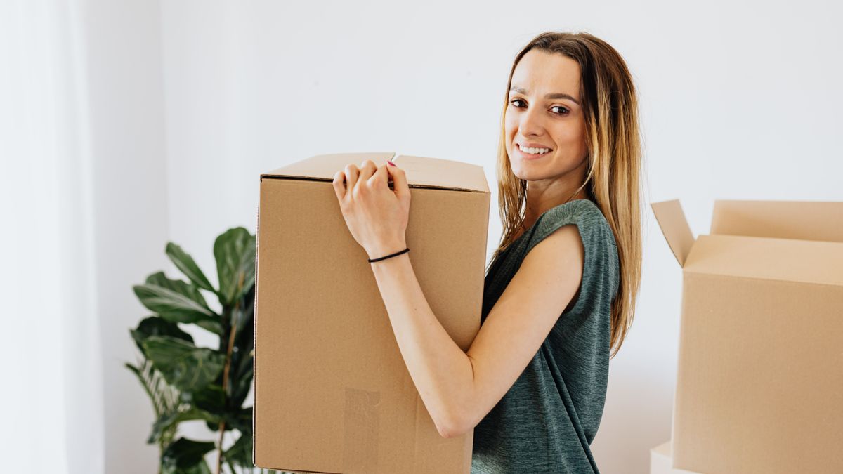 Side view smiling lady in casual green shirt carrying cardboard box while preparing for moving in new home and looking at camera happily