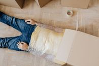 From above anonymous male in casual clothing lying on parquet with box on head and upper body wrapped in clingy plastic tape during moving out