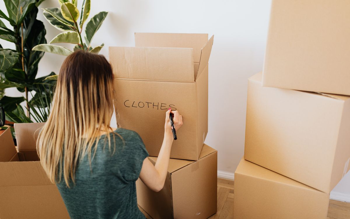 Back view faceless female in casual wear writing on cardboard container word clothes while packing belongings and preparing to move