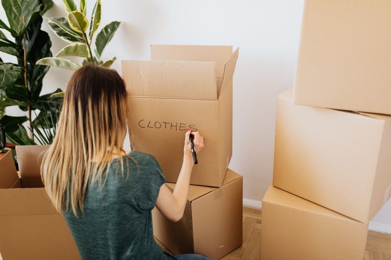 Back view faceless female in casual wear writing on cardboard container word clothes while packing belongings and preparing to move