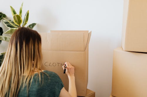 Back view anonymous female in casual outfit writing on cardboard container word cloth while packing belongings and preparing to move