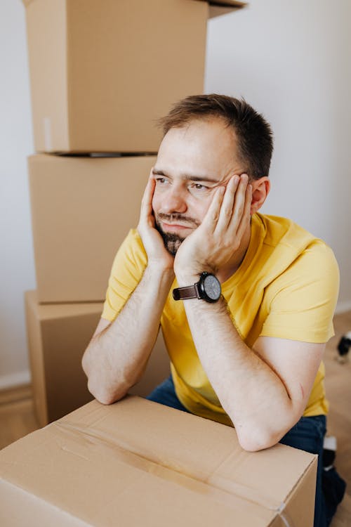 Frustrated man in casual wear holding face between hands while leaning on carton box and looking away with sadness after long tiring relocation day