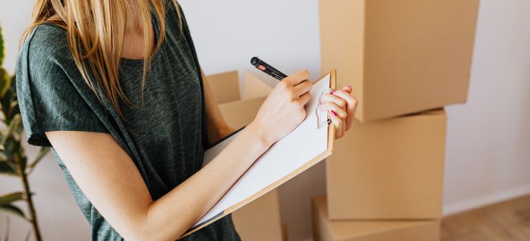 Woman checking a clipboard beside moving boxes while following tips for moving out for the first time.