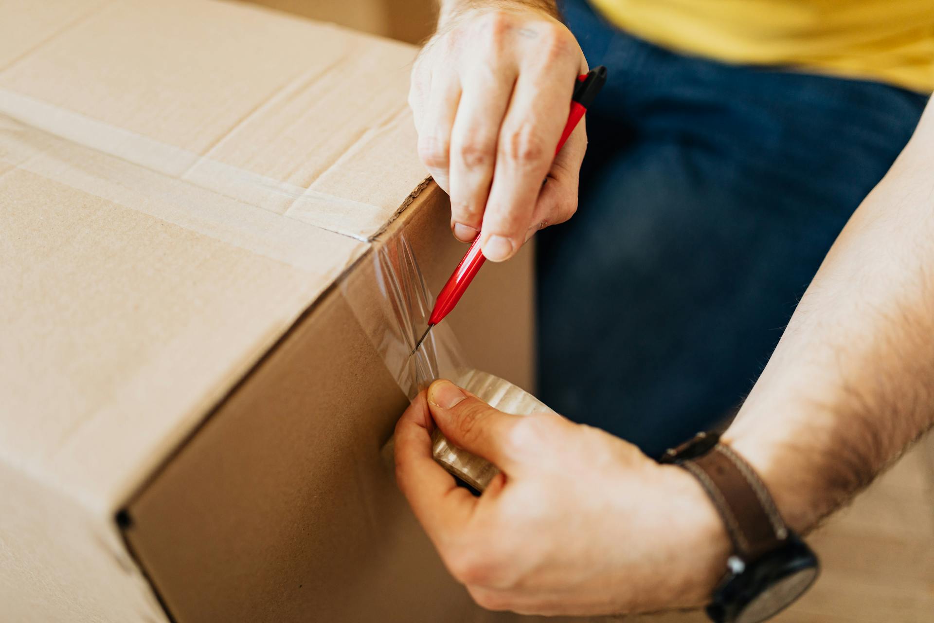 From above crop anonymous male sealing packed carton boxes by using tape and cutter knife while preparing for moving in new apartment