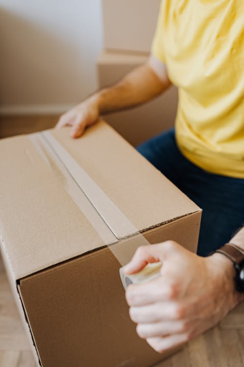 Crop anonymous guy in casual clothes using clingy tape to seal cardboard box for courier during relocation
