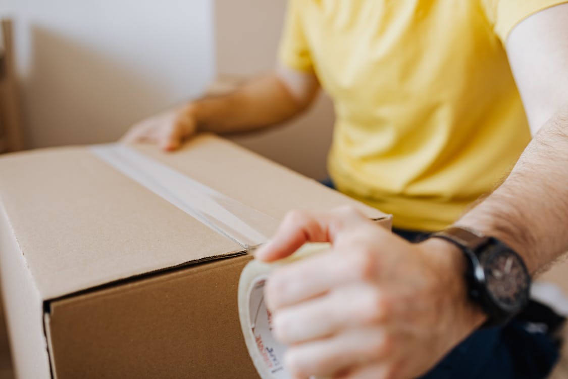 Free Crop anonymous male in casual wear using cling tape to seal cardboard container with belongings during relocation Stock Photo