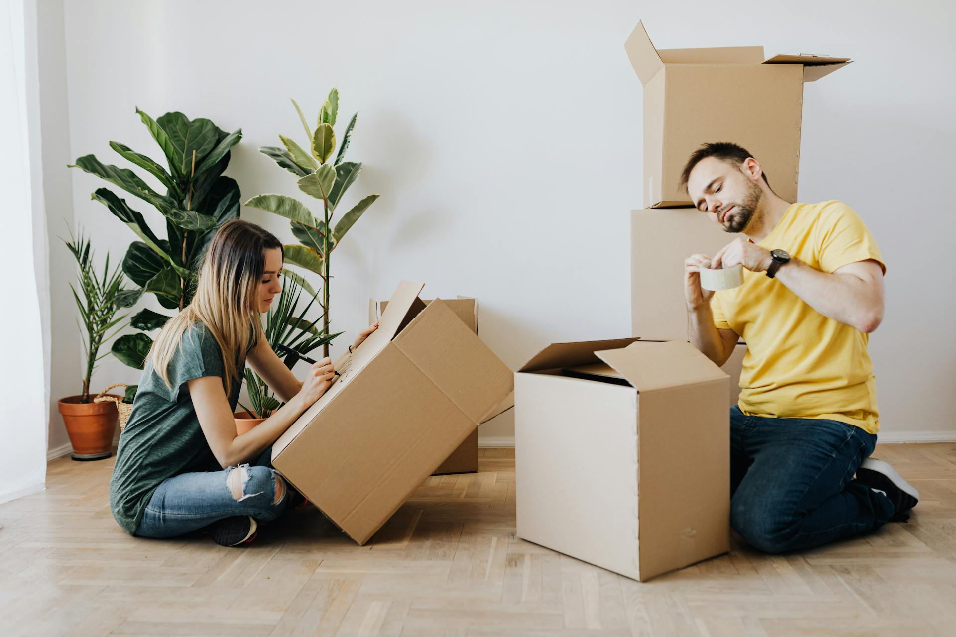 Side view full length glad couple packing stuff into cardboard boxes while sitting on floor at home during relocation in new house