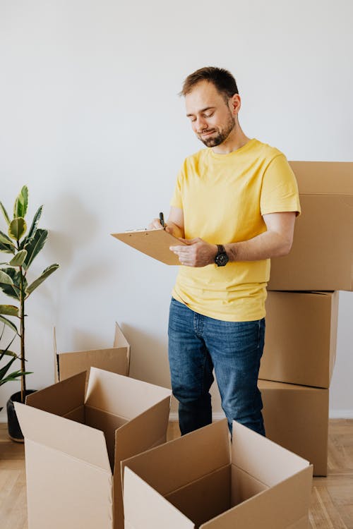 Free Full body content male in casual wear writing down notes in clipboard while pacing cardboard containers to relocate into new apartment Stock Photo