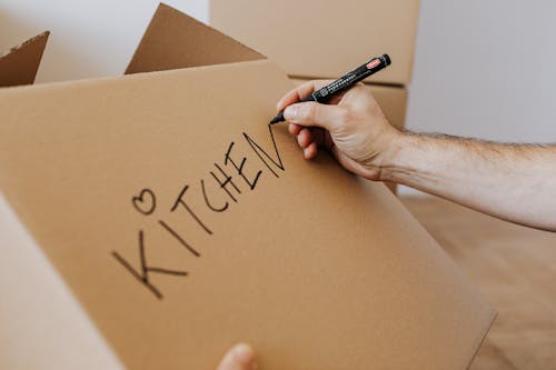 Crop unrecognizable male using marker to write on carton box word kitchen and draw cute heart while packing kitchenware before relocation