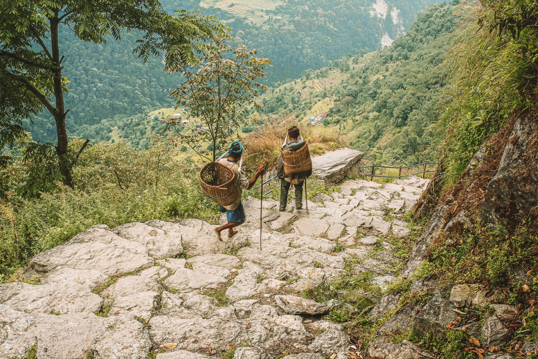 Two people carrying woven baskets hike down a stone path in Ghode Pani, Nepal.