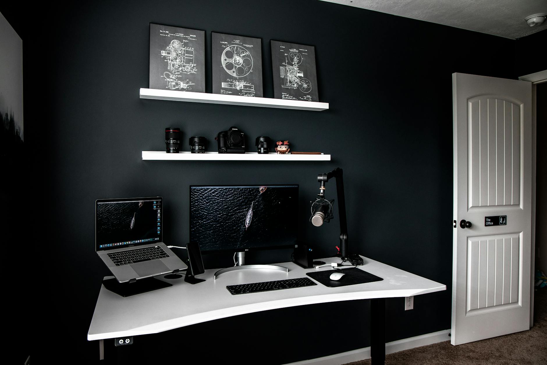 Modern style workspace with laptop near monitor and professional microphone on white table under shelves with schemes in frames