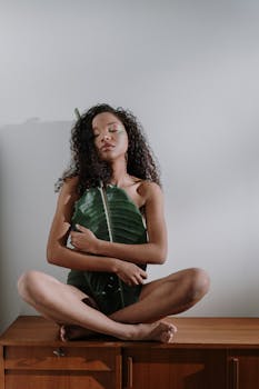 A woman meditates indoors, seated in lotus position with eyes closed, holding a large tropical leaf.