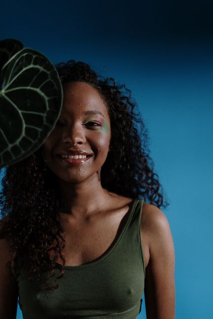 A joyful woman with curly hair and face art stands beside a green leaf, set against a blue background.