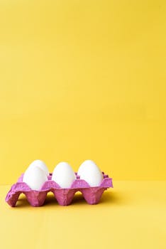Three white eggs in a pink carton against a vibrant yellow backdrop.