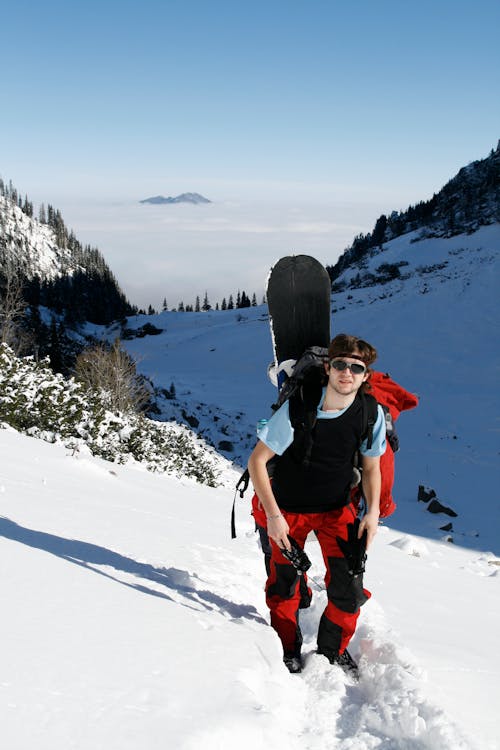 Full body male in outwear and sunglasses walking on rough terrain covered with snow in sunny day and looking at camera