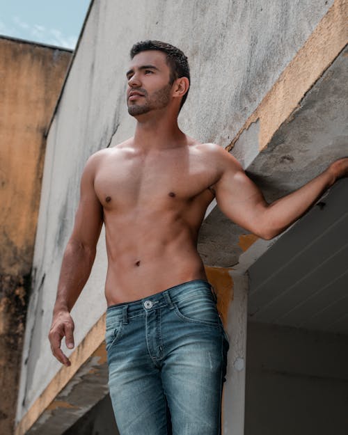 From below of serious ethnic shirtless male in jeans standing near shabby building in sunny day