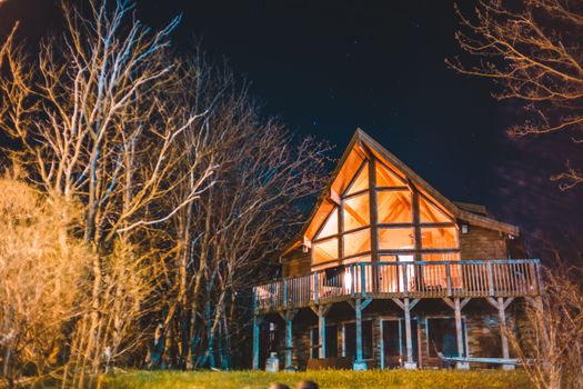 在星光灿烂的夜晚，现代小屋坐落在树木环绕的宽敞草地庭院中，外观带有装饰柱子和大阳台