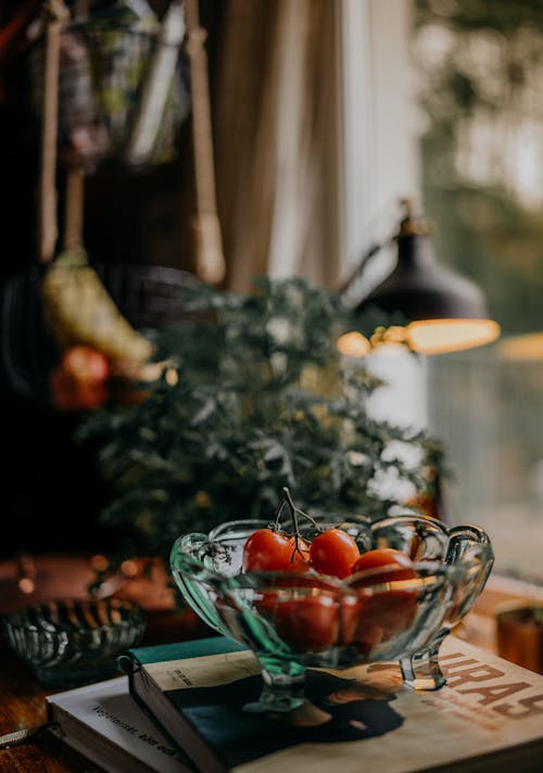 Ripe organic cherry tomatoes in glass bowl placed on stacked books on table near houseplant and lamp