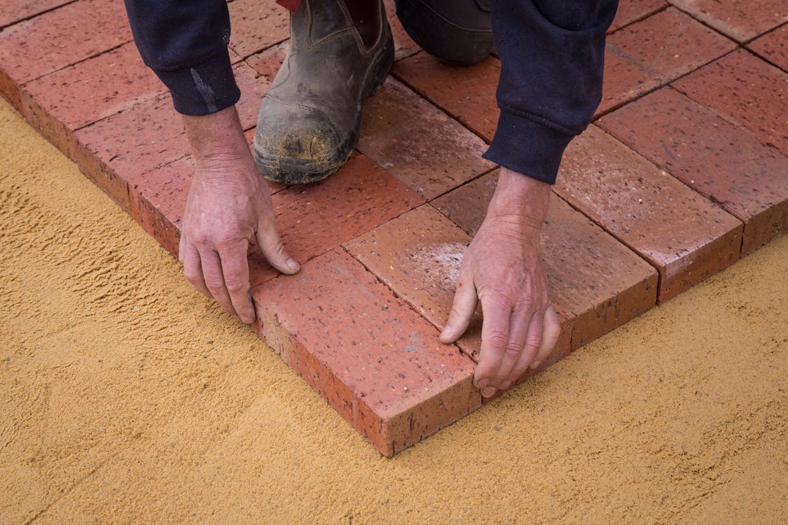 High angle crop anonymous worker in workwear lying bricks on sandy ground in square shape