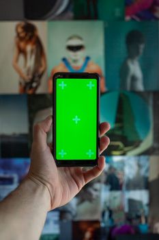 A hand holding a smartphone with a green chroma key screen in front of a collage wall.