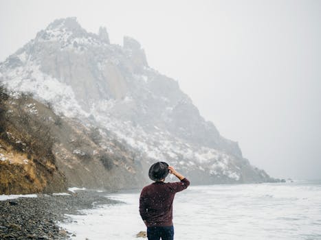 Person enjoying a snowy winter coast adventure, gazing at the dramatic rocky shoreline.
