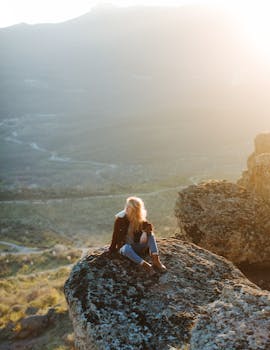 A serene outdoor scene with a woman enjoying the sunset atop a mountain rock.