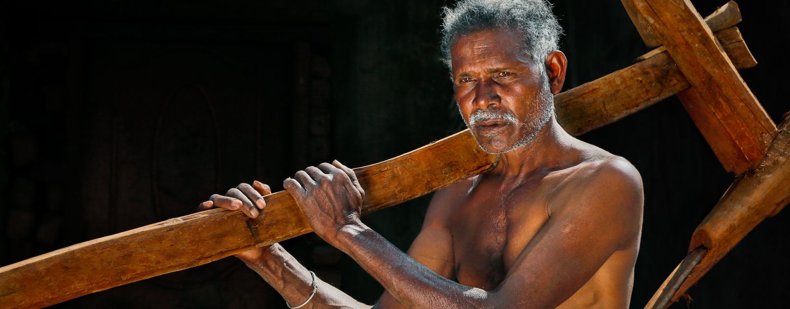 A shirtless elderly South Asian farmer standing with a wooden plow in Hanuman Junction, India.
