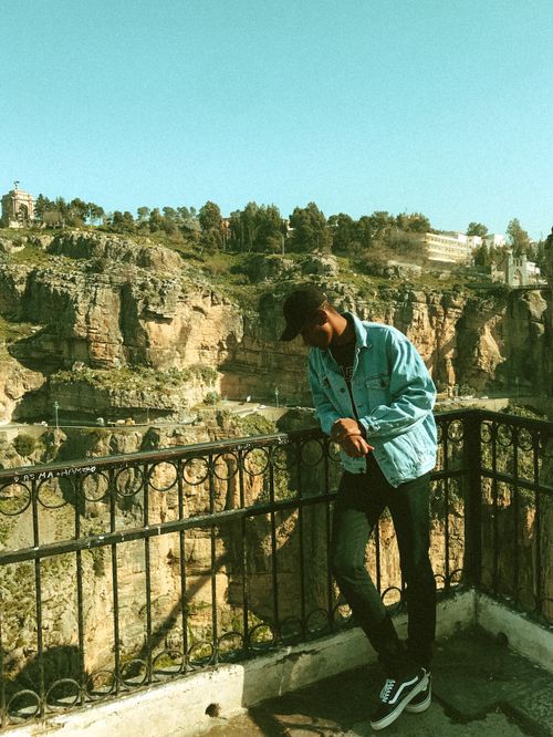 Free Full body of young ethnic male tourist in trendy outfit and cap leaning on metal railing of observation deck and relaxing in rocky terrain Stock Photo