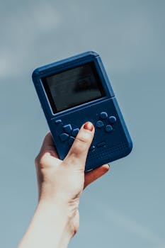 A close-up of a hand holding a vintage blue gaming console against a clear sky.