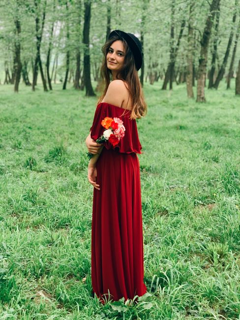 A smiling woman in a red dress stands in a lush green forest holding a bouquet.