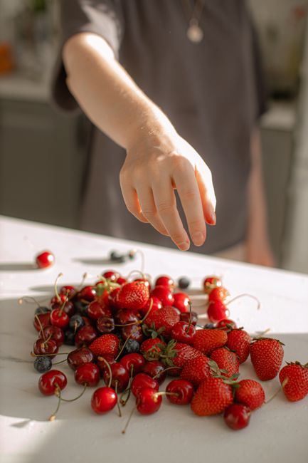 A hand reaches for fresh strawberries, cherries, and blueberries on a white table, bathed in sunlight.