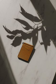一个时尚的皮革钱包放在大理石台面上，投下戏剧性的树叶阴影