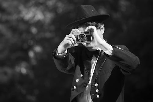 Grayscale Photo of Man in Black Hat and Coat Holding Camera