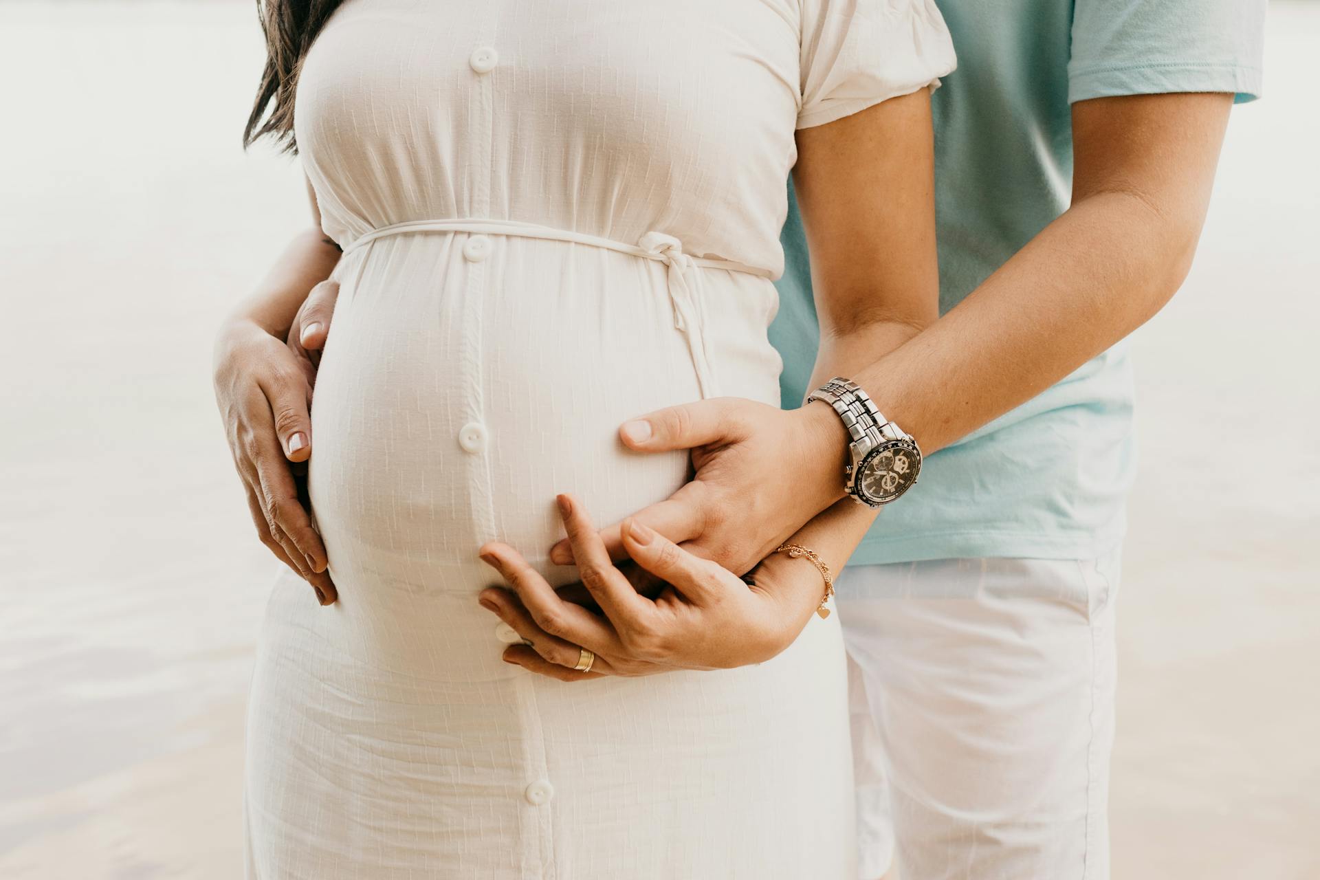 Crop anonymous young man in casual clothes embracing belly of pregnant wife while standing on seashore