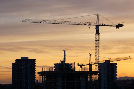 Silhouette of construction cranes and buildings against a vibrant sunset urban skyline.