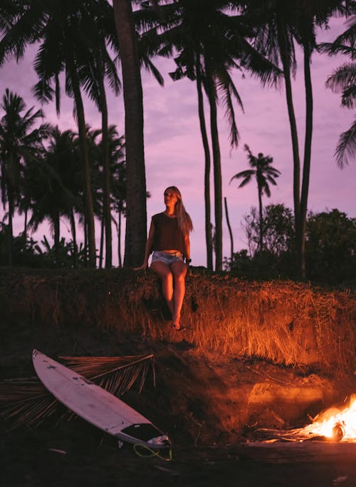 Full body of unrecognizable young woman relaxing on rocky ground near bonfire and surfboard against palm trees during amazing purple sunset